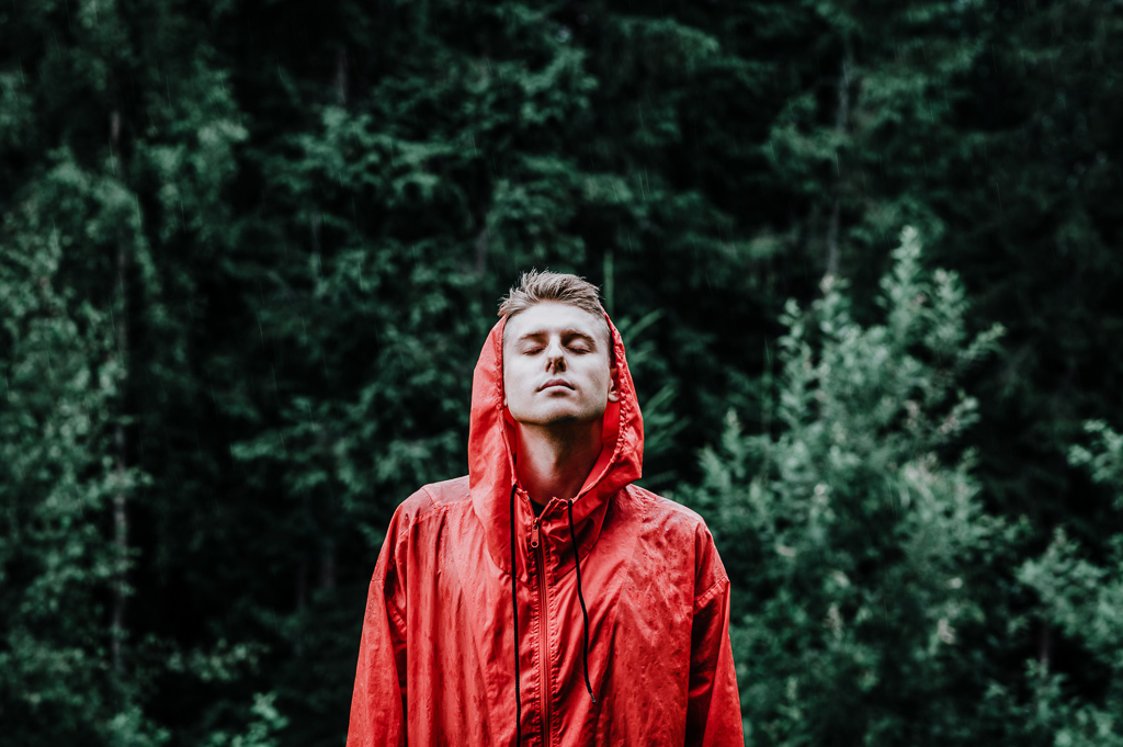 Handsome young man in a red raincoat stands outdoors in the rain with his eyes closed and enjoying the rain. Portrait of a young male hiker on a hike standing in the rain on a green background.